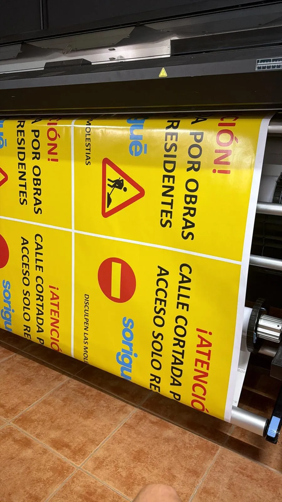 Rollo de vinilo saliendo de un plóter de gran formato con carteles amarillos de aviso de obras impresos